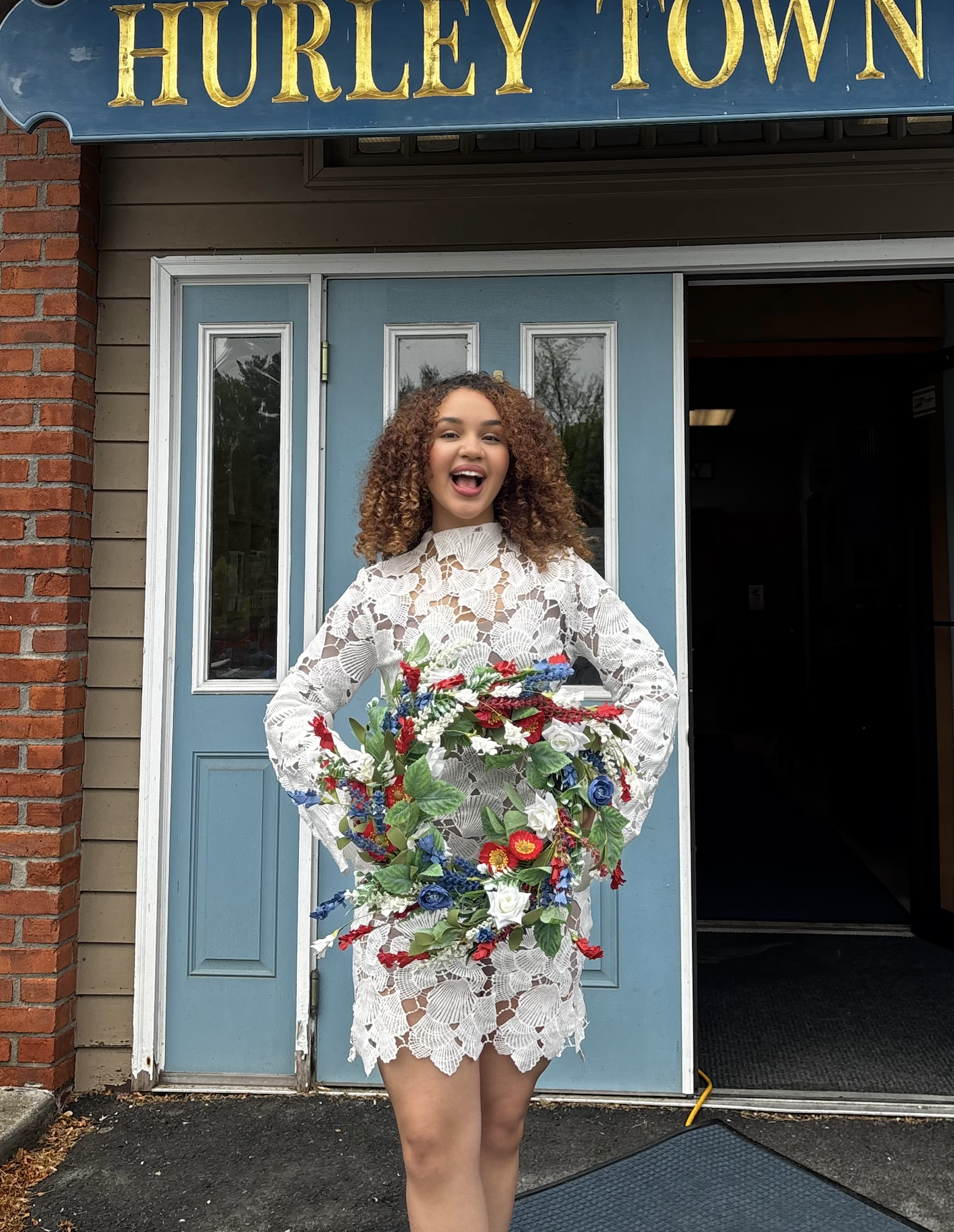 Mary Bellakbira in a white lace dress holding flowers at Hurley Town appearance as Miss Heart of New York