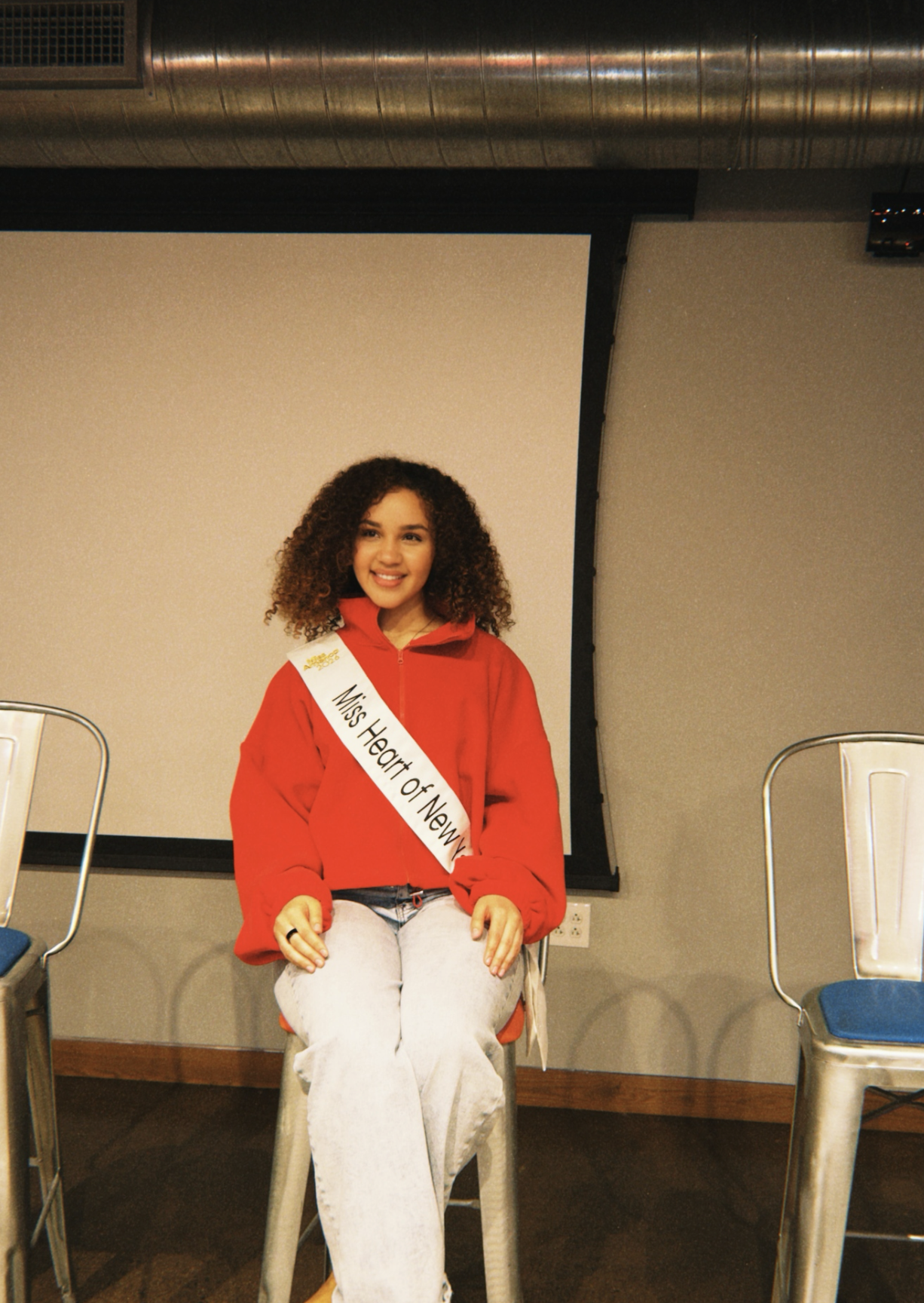 Mary Bellakbira in a red hoodie wearing her Miss Heart of New York sash, sitting on a stool at a New York appearance