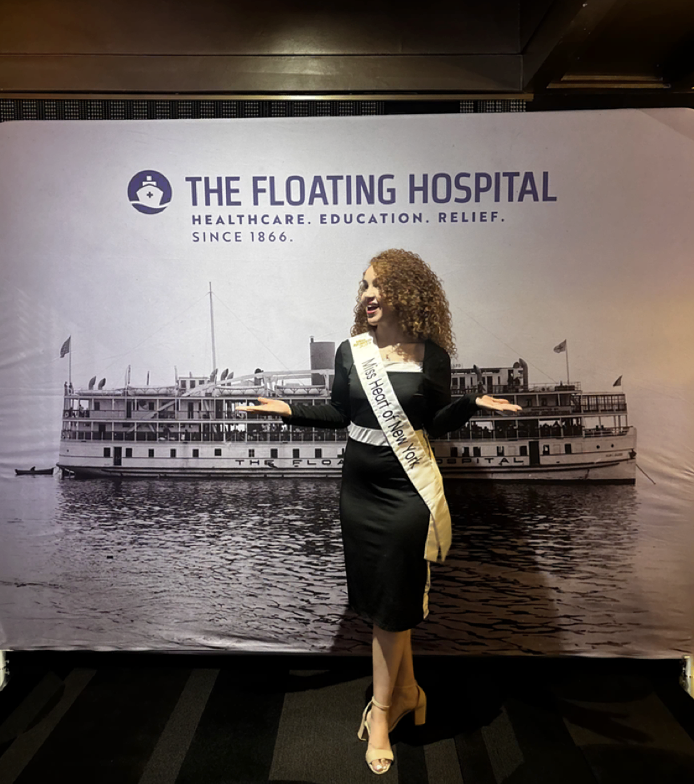 Mary Bellakbira in a black dress wearing her Miss Heart of New York sash in front of The Floating Hospital backdrop