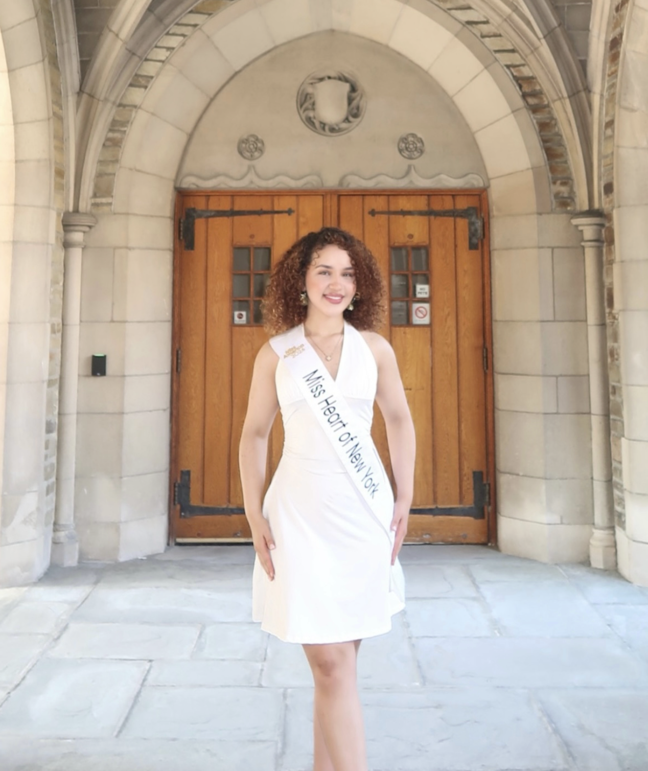 Mary Bellakbira in a white dress wearing her Miss Heart of New York sash standing in front of a stone archway